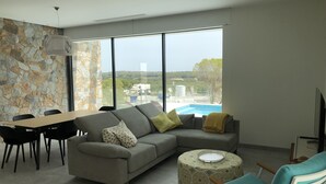 Un salon moderne avec un canapé gris, une table à manger avec des chaises, un pouf à motifs et une vue sur une piscine et des immeubles.