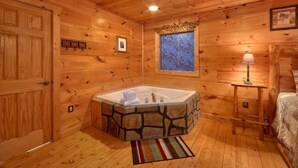 Ein Blockhausszimmer mit freistehender Steinbadewanne, Holzwänden, einem Fenster mit blauem Mosaik, einer Tür und einer Nachttischlampe.