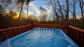 Ein Whirlpool, umgeben von einer Holzterrasse und Bäumen, mit einem Sonnenuntergang im Hintergrund.