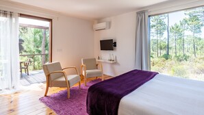 Une chambre avec un grand lit, deux fauteuils, une télévision et un balcon donnant sur la forêt.