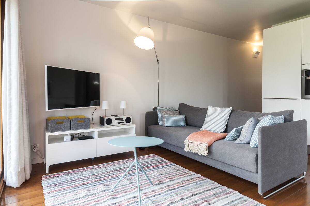 A modern living room with a grey sofa, a small round table, a flat-screen TV, a radio, and a floor lamp.