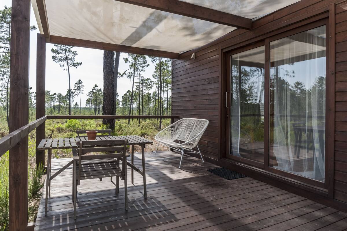 A wooden deck with a table and chairs, a covered area, and a glass door leading to a garden.