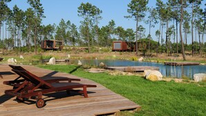 Une terrasse en bois avec des chaises longues, un petit étang et des cabanes situées dans une zone boisée.