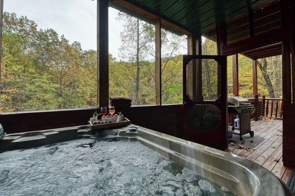A hot tub with a wooden deck, a grill, and a view of autumn trees.