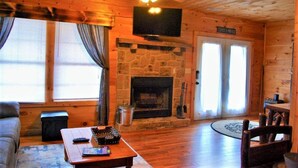 A cozy log cabin interior with a stone fireplace, wooden floors, a ceiling fan, and a television mounted above the fireplace.