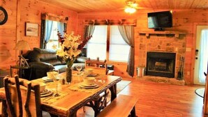 A cozy cabin interior with a wooden dining table set for four, a fireplace, a television mounted above it, and a leather sofa in the background.