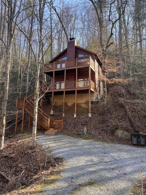 A two-story cabin with a balcony, surrounded by a forested area.
