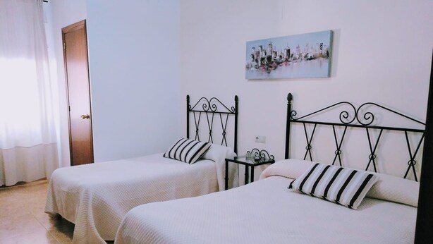 A room with two beds, each with a black metal headboard and white bedding. There is a small table with a lamp between the beds, and a painting on the wall above the beds. - Pensión Cuadrado (Seron)