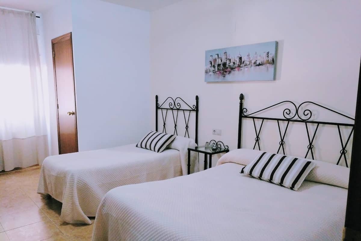 A room with two beds, each with a black metal headboard and white bedding. There is a small table with a lamp between the beds, and a painting on the wall above the beds.