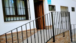 A building with a white exterior, a wooden door, and a black metal staircase leading to a stone-paved area. - Pensión Cuadrado (Seron)