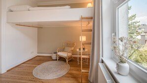 A compact room with a loft bed, a small chair, a table, and a window with a view of buildings.