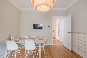 Un coin repas avec une table en bois, des chaises blanches, un téléviseur et un mur décoré de motifs.