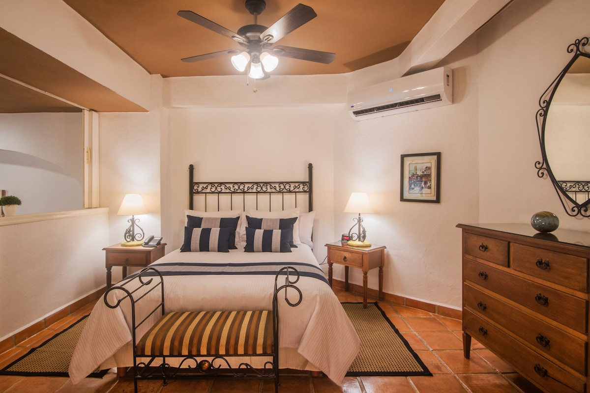 A neatly arranged bedroom with a bed, two bedside tables with lamps, a dresser, a ceiling fan, and an air conditioning unit.