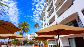 Zona de piscina con sillones y sombrillas, cielo azul despejado y un edificio blanco con balcones.