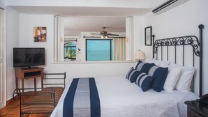 Habitación de hotel con cama, televisor, ventilador de techo, aire acondicionado, ventana con vista al océano y una pequeña mesa con lámpara.