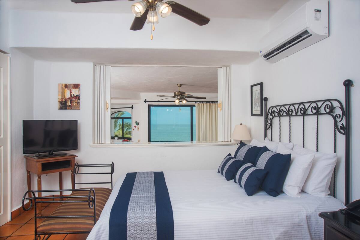 A hotel room with a bed, a television, a ceiling fan, an air conditioner, a window with a view of the ocean, and a small table with a lamp.