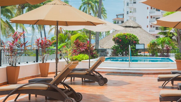 Zona de piscina con sillones y sombrillas, rodeada de plantas tropicales y vistas a un edificio con balcones.