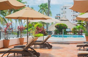 A poolside area with lounge chairs and umbrellas, surrounded by tropical plants and a view of a building with balconies. - One Beach Street Zona Romantica Puerto Vallarta (Puerto Vallarta)