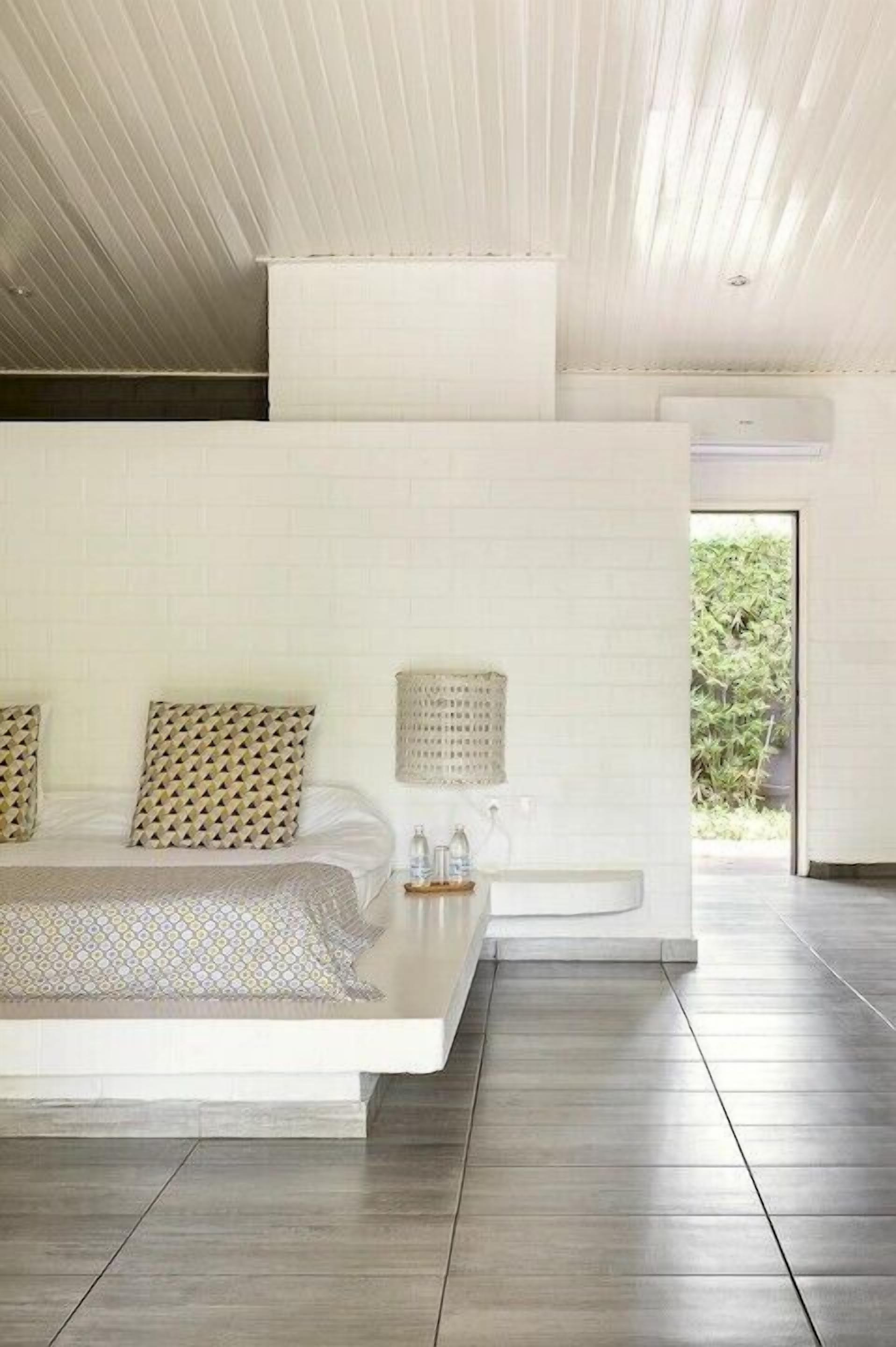 A modern bedroom with a large bed, a bedside table with a lamp, and a view of greenery outside.
