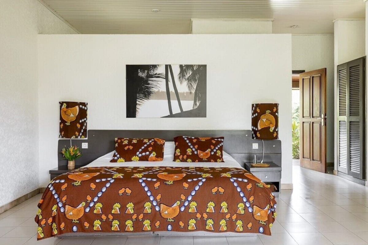 A bedroom with a large bed, a nightstand, a wall-mounted artwork, and a view of the outdoors.