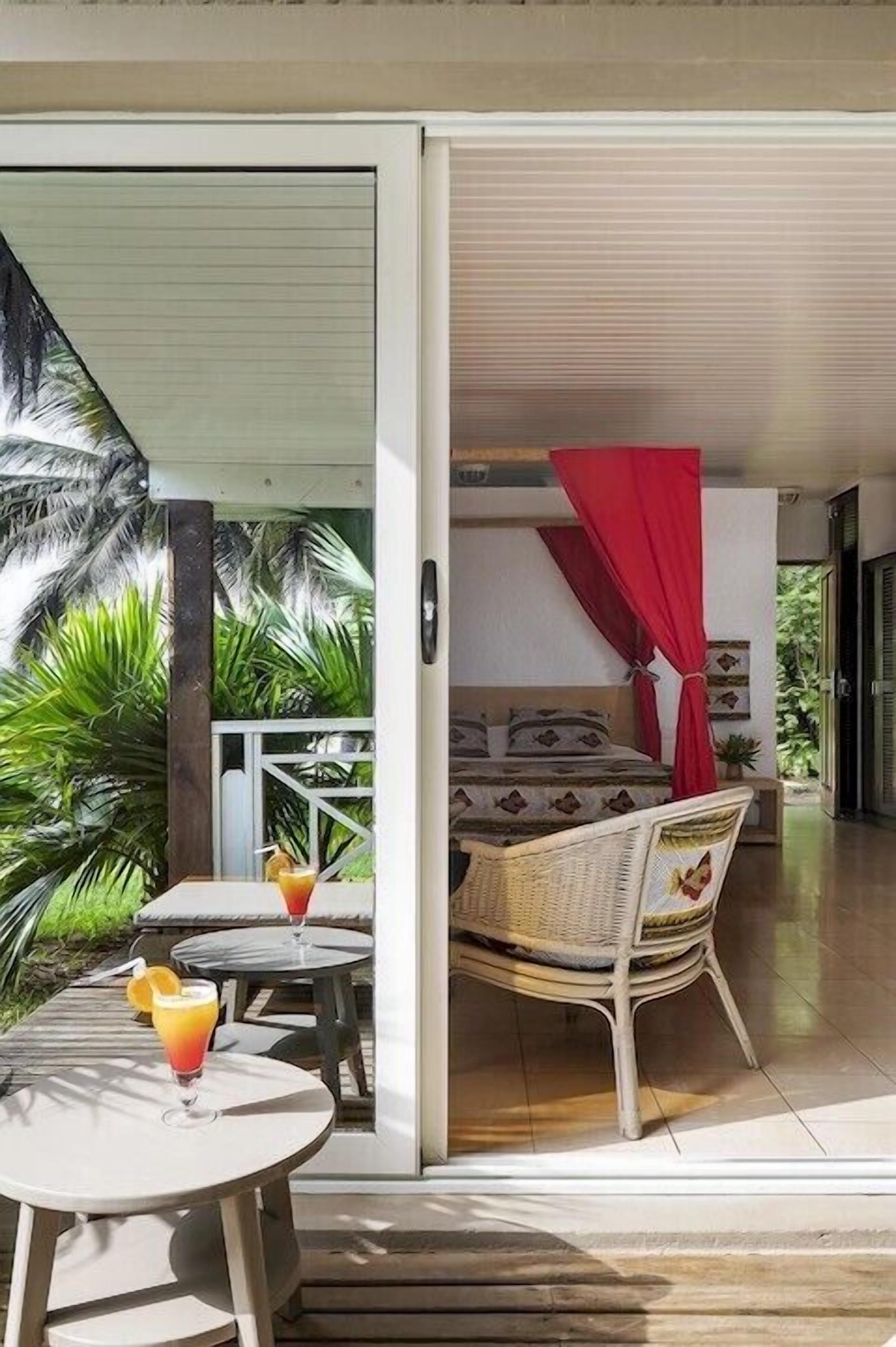 Una habitación con cama, dosel rojo, una silla de mimbre y una mesa con bebidas.