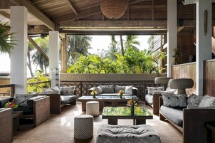A covered outdoor seating area with multiple sofas and tables, surrounded by lush greenery and palm trees.