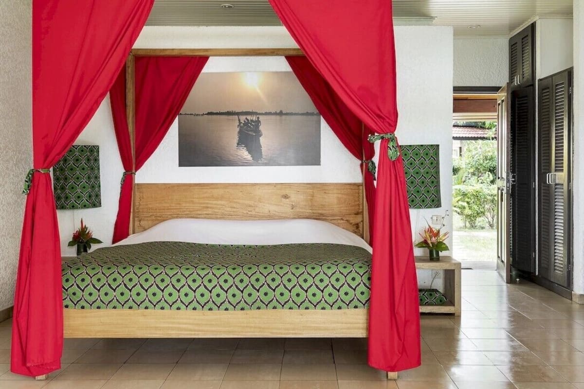 A bed with a green patterned bedspread, red curtains, and a framed picture of a sunset over water.