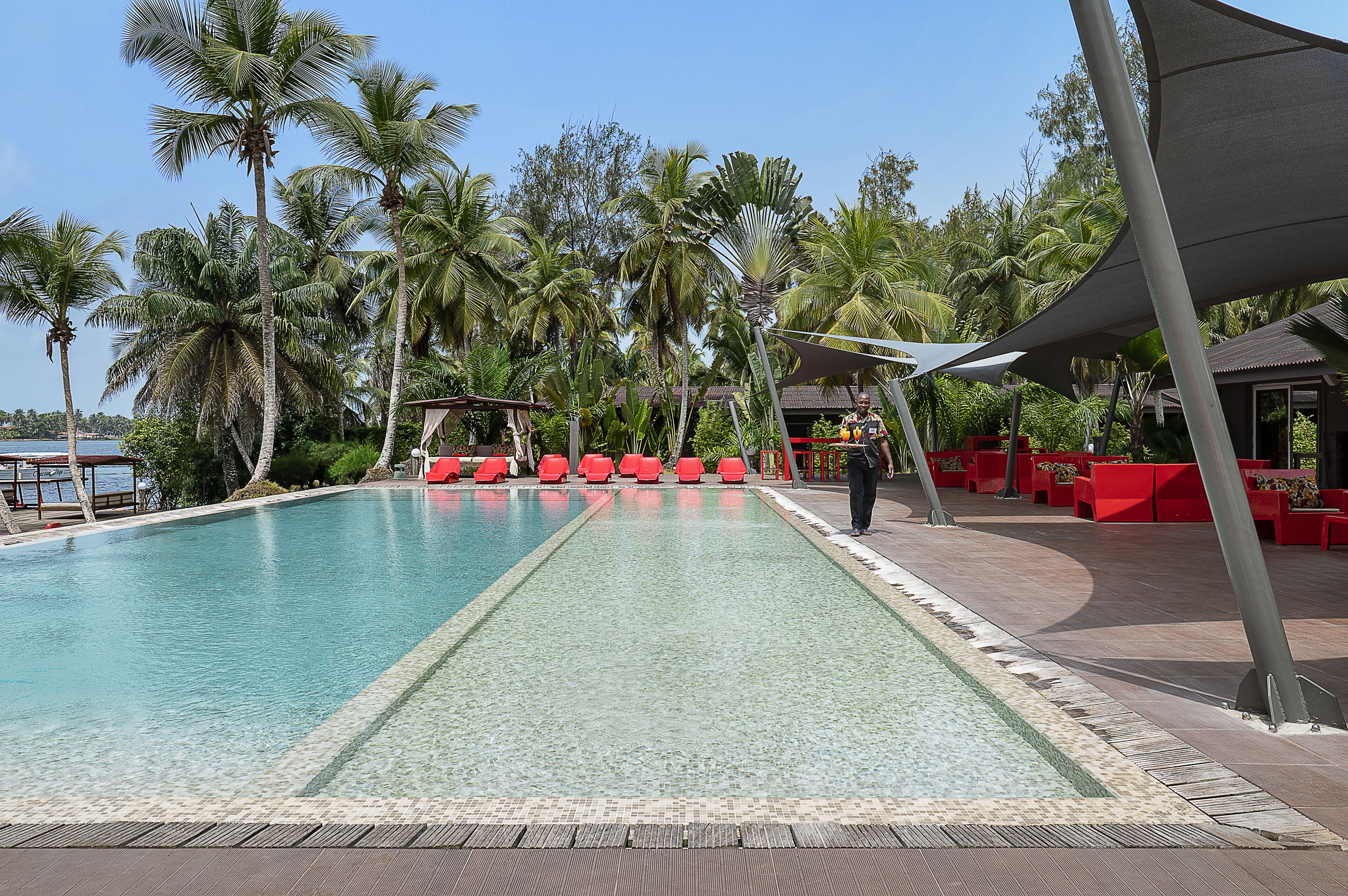 Piscina con sillas rojas y una zona sombreada, rodeada de palmeras.