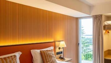 A hotel room with two beds, a nightstand with a lamp, and a view of greenery through the window.