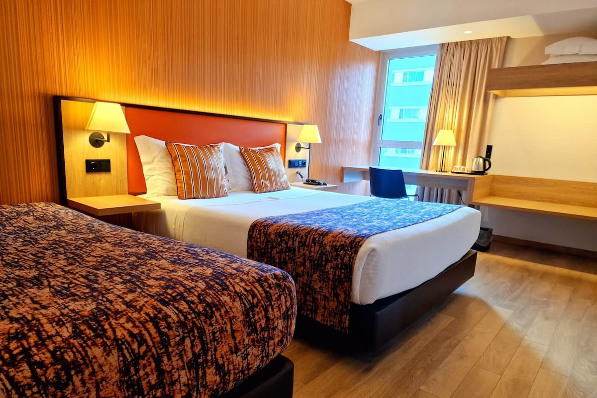 Ett hotellrum med två sängar, sänggavlar i trä, sänglampor, ett litet bord, en stol och ett fönster med gardiner.