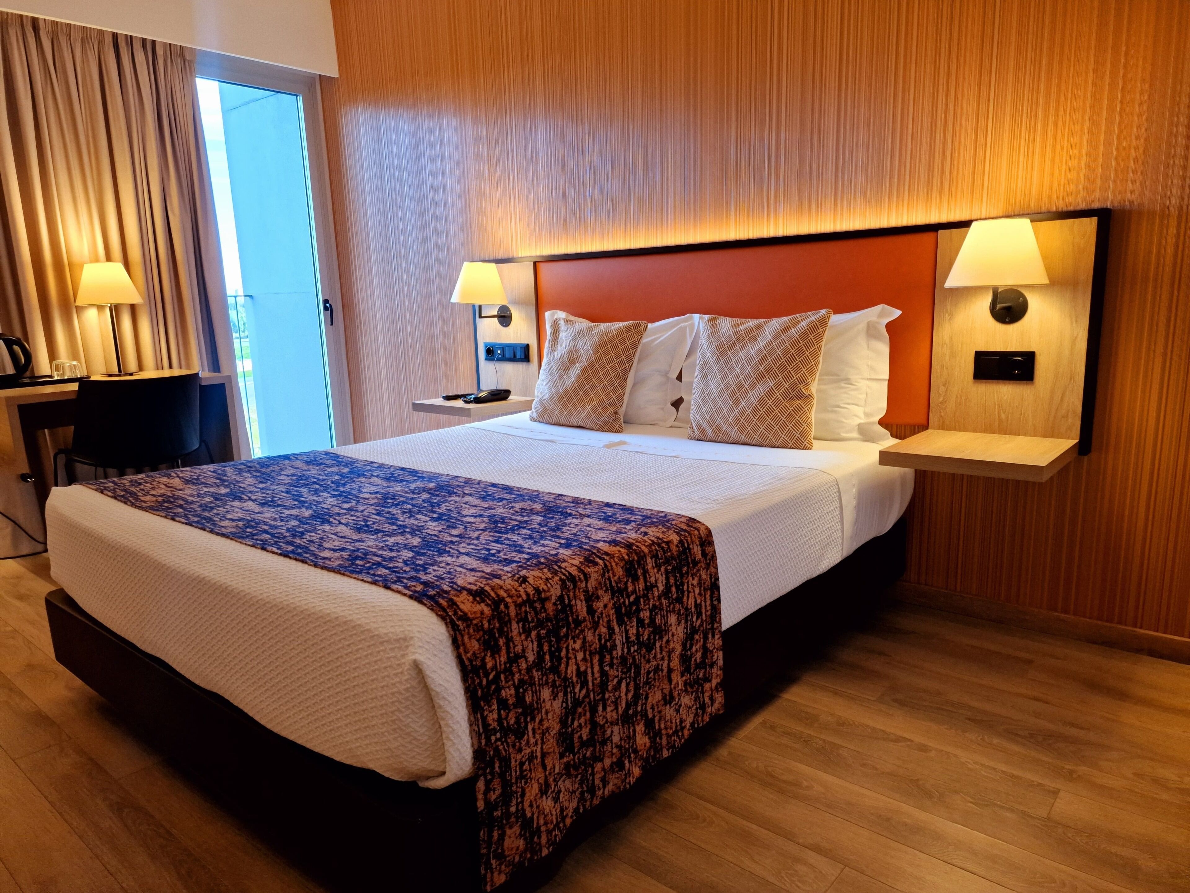 A hotel room with a bed, bedside lamps, a desk with a lamp, and a view of the outdoors.