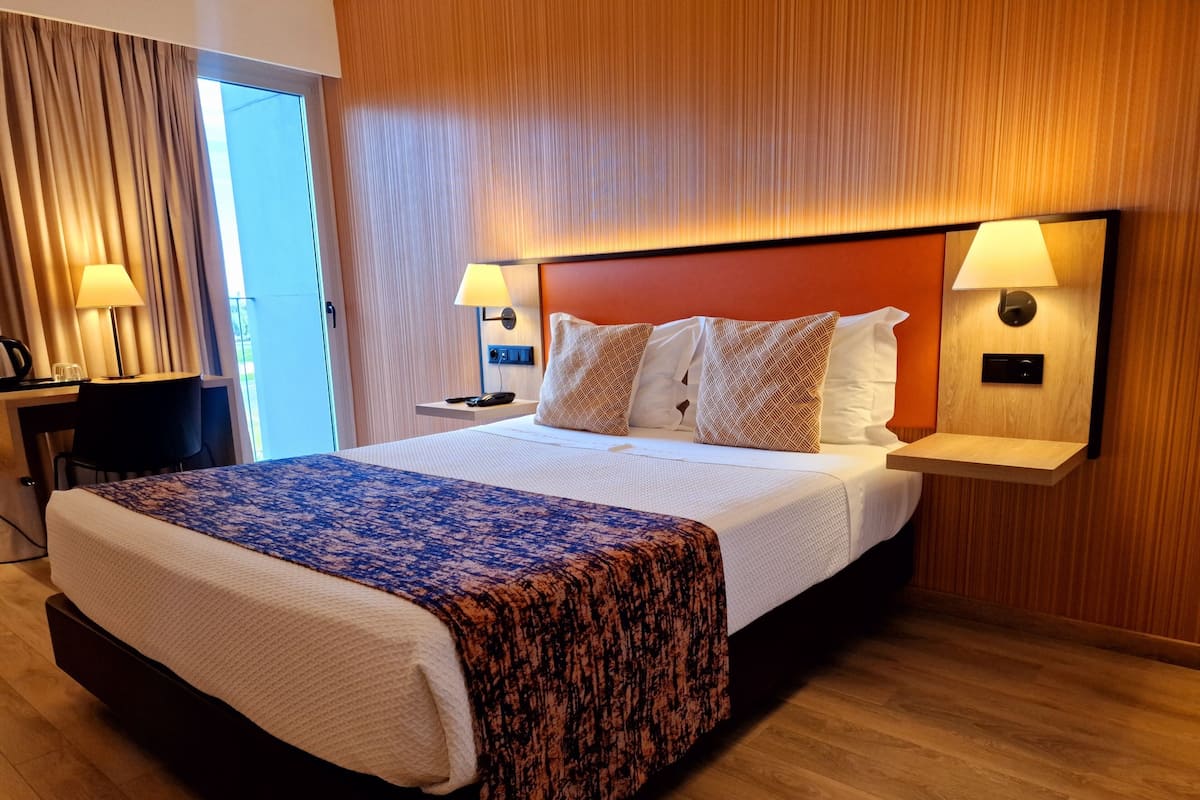 Ett hotellrum med en stor säng, två sänglampor, ett nattduksbord, ett skrivbord med en lampa och ett fönster med gardiner.