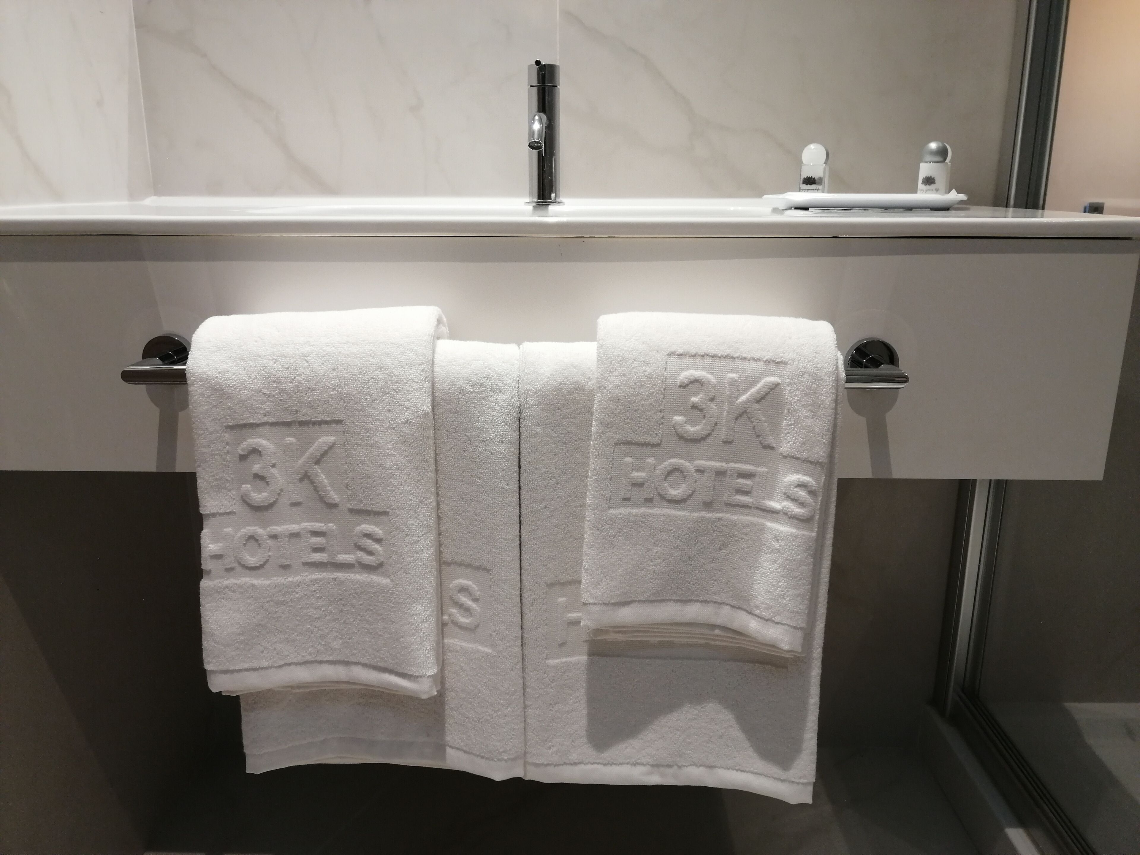 A bathroom with a white marble countertop, a modern faucet, and two towels hanging on a towel rack with "3K HOTELS" embossed on them.