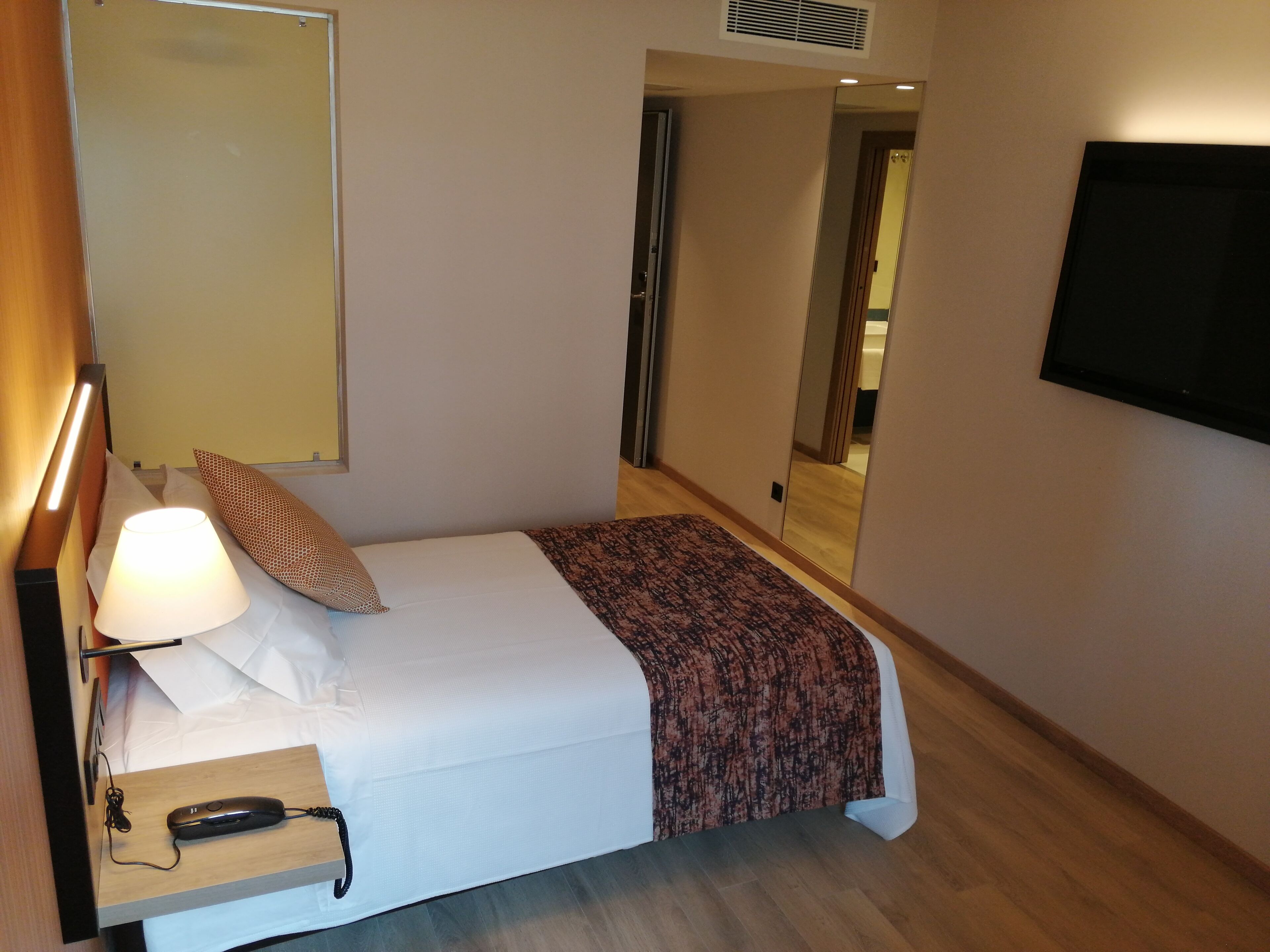 A hotel room with a bed, a bedside table with a lamp, a telephone, a mirror, and a television mounted on the wall.