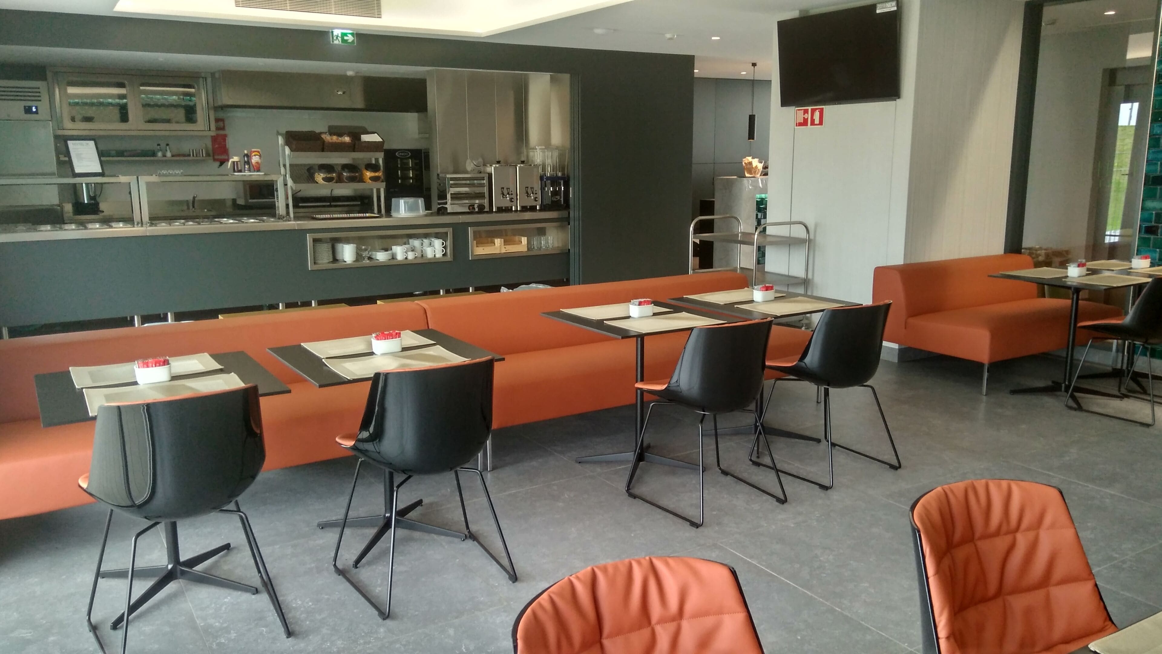 Een modern restaurant met oranje zitplaatsen, zwarte stoelen en een toonbank met keukenapparatuur.
