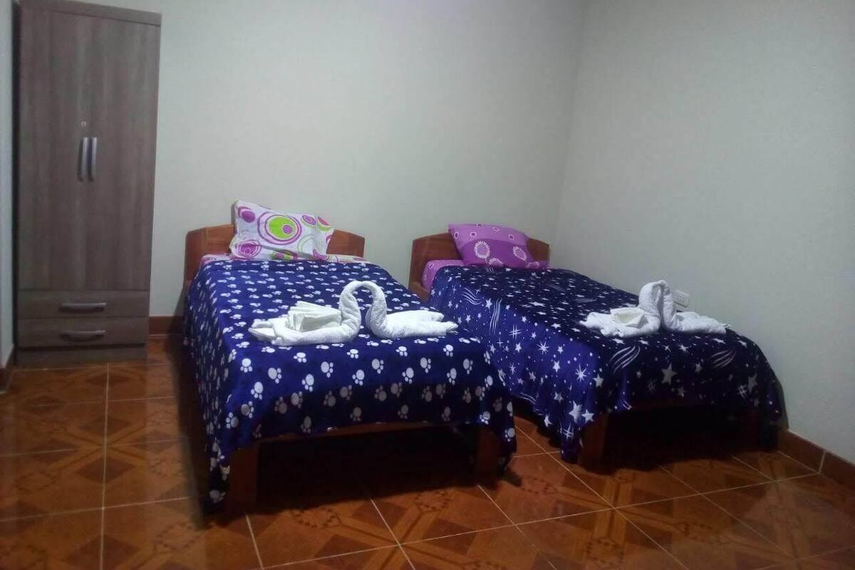 A room with two single beds, each with a blue bedspread and white pillows. There is a wardrobe on the left and a small table with a lamp.