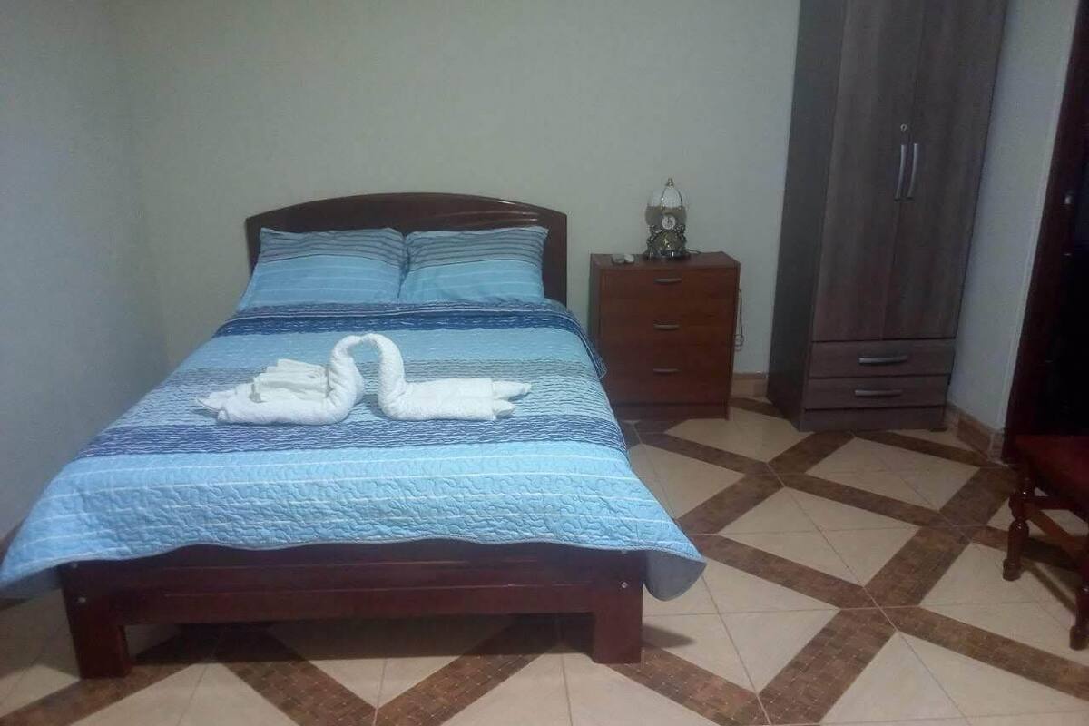 A bed with a blue quilt and two white swan-shaped pillows, a wooden nightstand with a lamp, and a wardrobe in a room with patterned tile flooring.