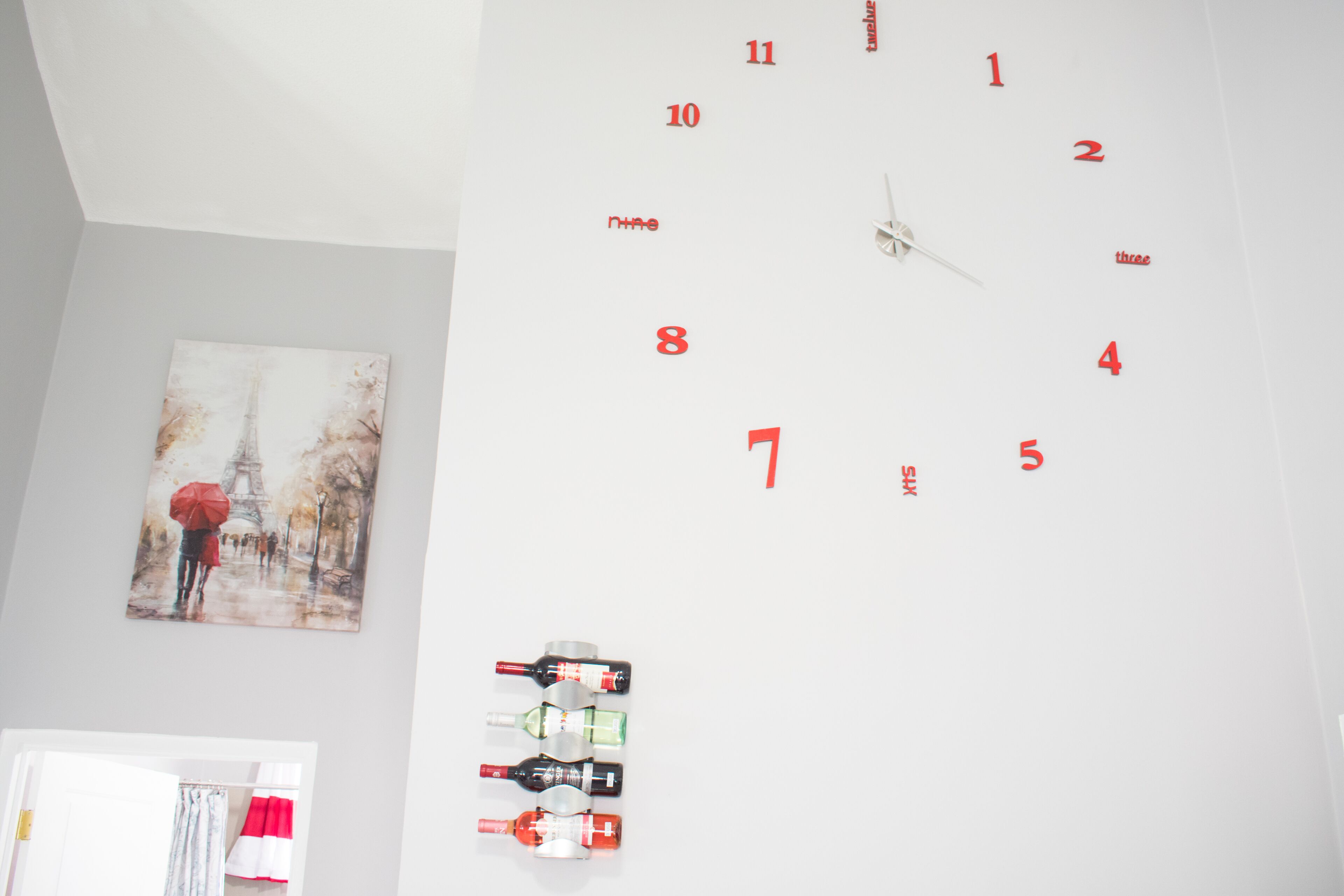A wall clock with red numbers and hands, a painting of the Eiffel Tower, and a row of wine bottles.