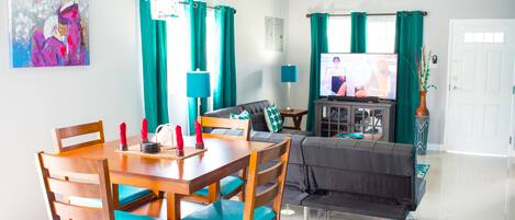 A modern living room with a dining area, teal curtains, a flat-screen TV, and a painting on the wall.