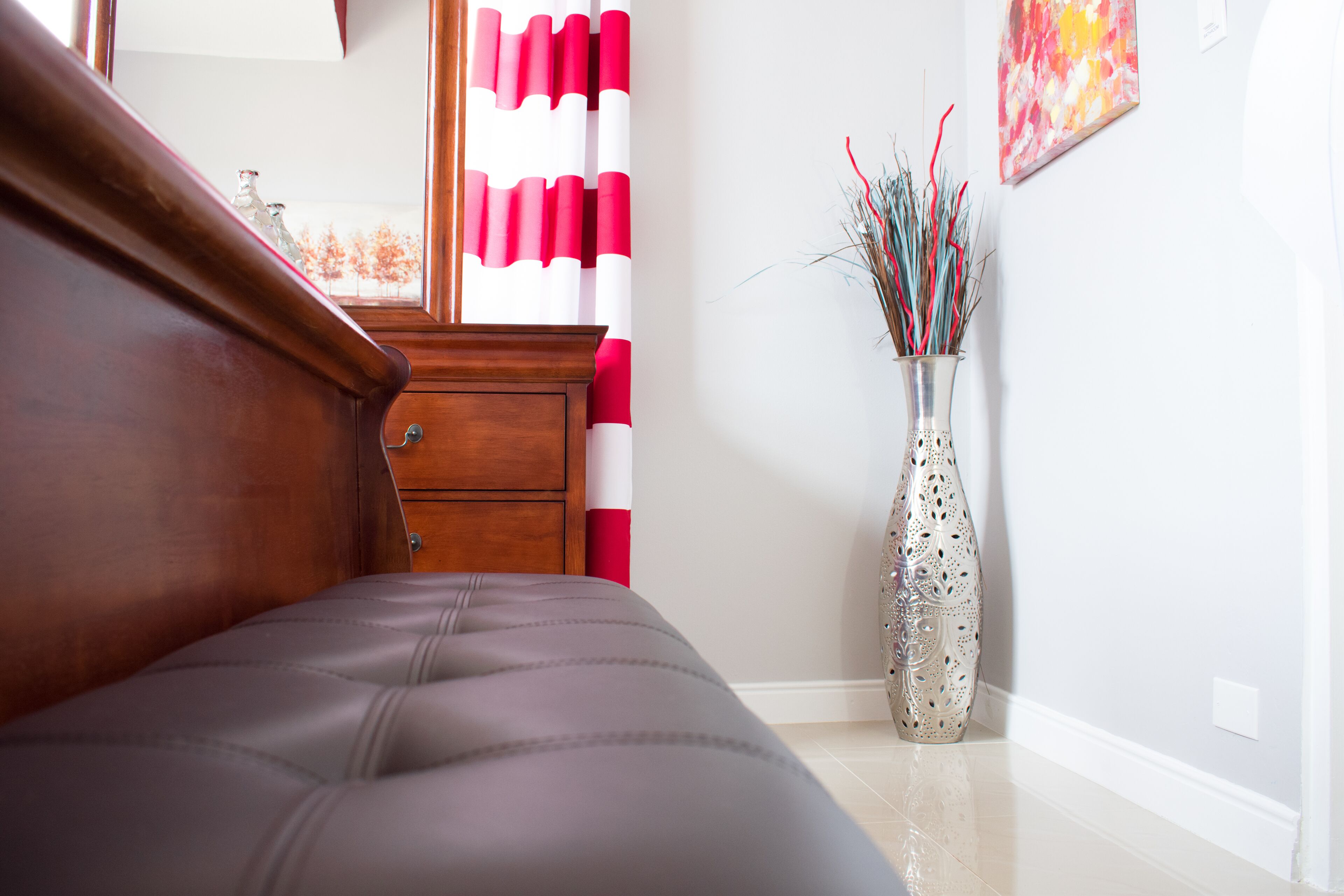 Une chambre à coucher avec une tête de lit en bois, un lit recouvert d’un tissu gris, un rideau à rayures rouges et blanches, et un vase décoratif orné de branches.