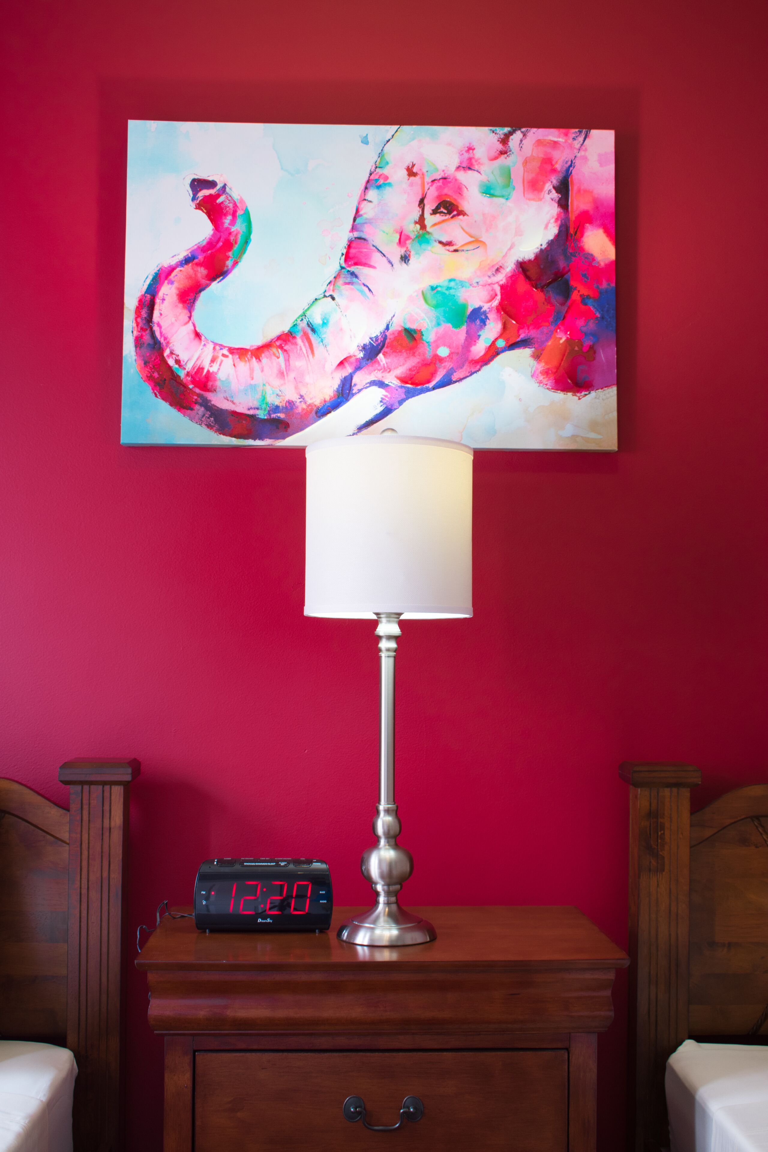 A room with a red wall, a painting of an elephant, a bedside table with a lamp, and a digital clock.