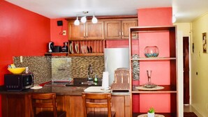 A compact kitchen with a red wall, wooden cabinets, a microwave, and a small dining area with two chairs. - New Kingston Guest Apartment At Chelsea (Kingston)