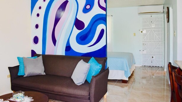 A living room with a brown sofa, a large abstract painting, a bed with a blue cover, and a white door with a geometric design.