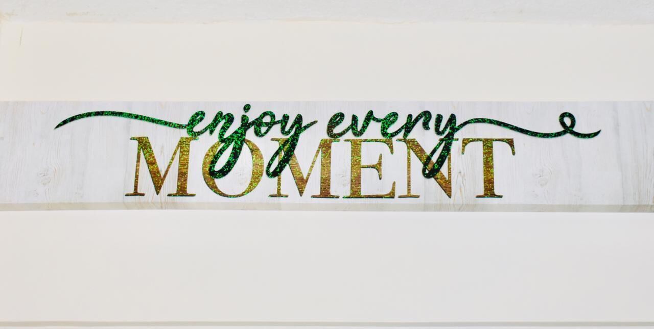 A sign with the text "enjoy every MOMENT" in glittery gold letters on a white background.