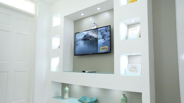 A modern living room with a flat-screen TV mounted on a wall, surrounded by glass shelves with decorative items. - Kingston Guest Apartment At Valencia (Kingston)