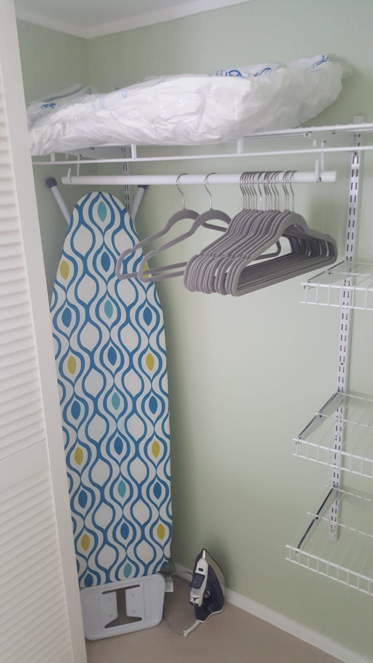 A closet with hanging clothes, an ironing board, and a shelf unit.