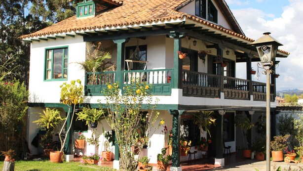 Una casa de dos pisos con balcón, rodeada de macetas y un jardín.