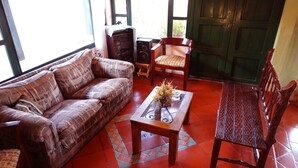 Un salón acogedor con un sofá estampado, una mesa de centro de madera con un jarrón de flores, una silla de madera y una puerta verde al fondo.