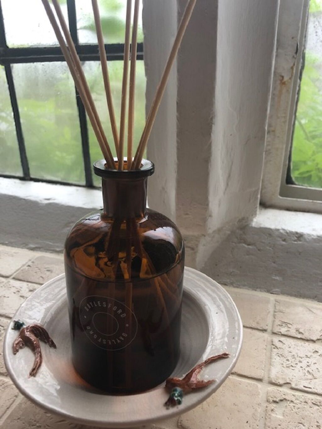 A dark brown bottle with a label, placed on a white tray with a decorative item beside it, in front of a window with a view of greenery.