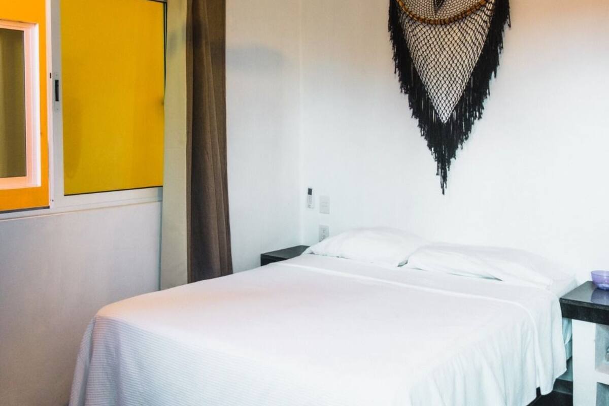 Et hotelværelse med en seng, ventilator, et billedværk på væggen og et vindue med gardiner.